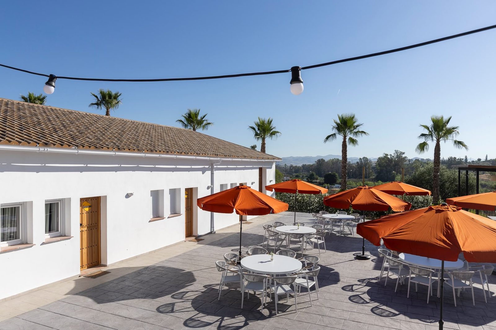 Het gedeelde terras voor de appartementen, met witte tafels, oranje parasols, lichtjes boven het terras en uitzicht op de palmen en de bergen